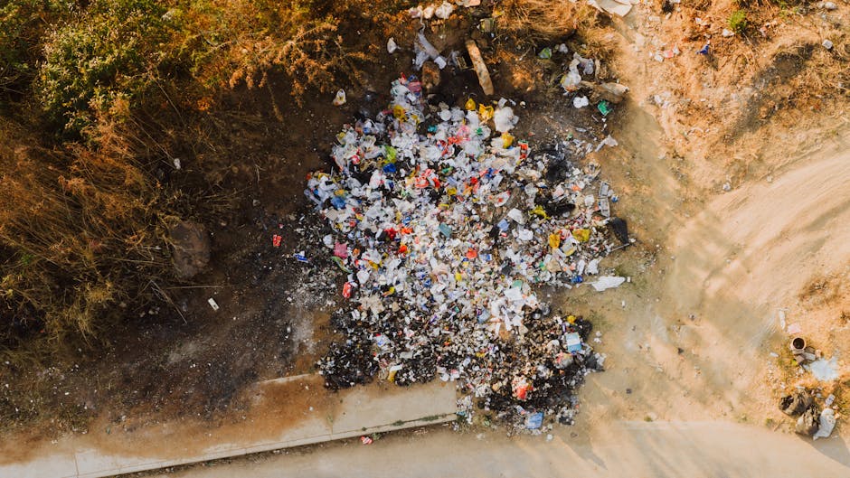 An aerial view of a small, unmanaged dump site on a patch of bare, brown soil adjacent to a concrete pavement and sparse vegetation, featuring a large heap of mixed waste including plastic bottles, wrappers, and other household rubbish. The waste appears scattered and loosely piled, with some items partially buried in the soil and others resting on the surface. The surrounding area shows signs of natural wear and erosion, with the edges of the heap blending into the surrounding earth, indicating a site for informal disposal of discarded household or commercial waste. There are no visible waste management equipment or containers in the image, implying it is an unmanaged or alternative waste collection point, which highlights the importance of professional rubbish removal services to prevent illegal dumping and promote environmental responsibility. The overall scene emphasizes the need for proper waste handling and disposal, especially in contexts where private collection or on-site clearance might be preferable to prevent environmental hazards and comply with local rubbish dumping laws.