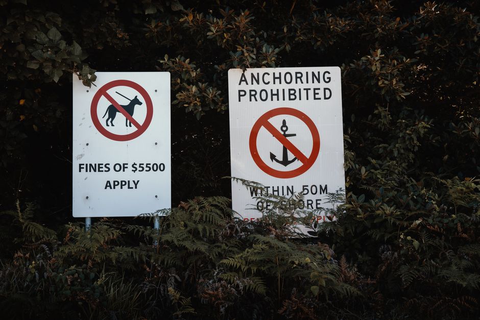 Two white outdoor warning signs are mounted on metal posts amidst dense foliage, with dark green and brown leaves partially obscuring the lower portions of the signs. The left sign displays a black silhouette of a dog with a red circle and slash crossing through it, indicating no dogs allowed, and includes the text 'FINES OF £5500 APPLY' below. The right sign features bold black text reading 'ANCHORING PROHIBITED' along with a symbol of an anchor crossed out within a red circle, and additional smaller text indicating the restriction applies 'WITHIN 50M OF.' The signs are situated on a natural, slightly overgrown area, suggesting they are meant to regulate activities in a coastal or waterfront setting, and relate to private or independent waste management and vessel mooring regulations in the context of rubbish removal or environmental protection.