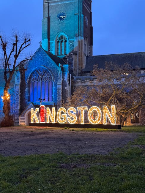 A historic church building with a tall clock tower constructed from stone and brick, featuring Gothic-style arched windows with stained glass. The church is illuminated by blue lighting that highlights its architectural details against a dusky evening sky. In front of the church, there is a large, brightly lit sign spelling 'KINGSTON' with each letter outlined in small light bulbs, positioned on a grassy area with patches of soil. To the right of the sign, a leafless tree with twisting branches extends outward, partially obscuring the church’s facade. The overall scene reflects an outdoor, urban environment in the late evening or early night, where the illuminated sign and lighting accentuate the setting. The image suggests a location that could be associated with a town or city centre, where local signage and landmark architecture are prominently displayed, supporting the context of community and place while subtly aligning with themes of private or independent urban waste handling, such as rubbish removal services. The lighting and environment create a clear visual contrast between the historic building and modern signage, situating the scene in a public space conducive to waste or rubbish collection activities by local providers like houseclearancekingstonuponthames.co.uk.