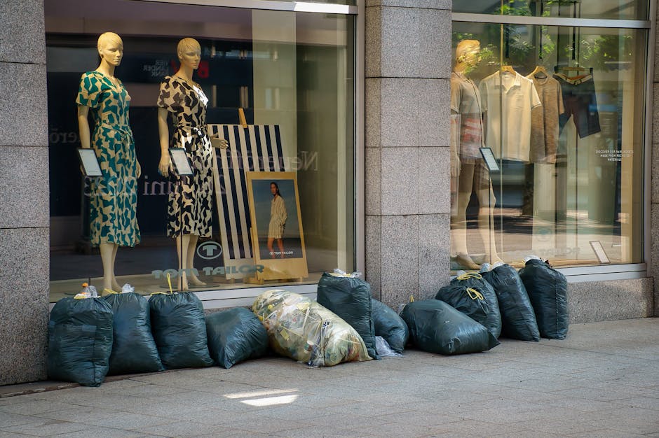 A large pile of mixed household rubbish and waste materials, including black and red trash bags, flattened cardboard boxes, crumpled paper, and discarded packaging, is overflowing from a grey communal waste bin situated on a paved sidewalk area. The litter surrounds the bin on the ground, with some debris spilling onto the nearby brick paving. Behind the waste collection point, there are parked vehicles, including a silver car with a yellow registration plate, and behind that, a row of shops with brightly coloured signage and a prominent yellow storefront. To the left of the rubbish, a metal railing and a tree trunk are visible, with the background showing a building under scaffolding and construction netting, indicating possible maintenance or renovations. The scene appears to be during daylight, with natural lighting casting soft shadows, illustrating an example of private waste handling or alternative rubbish collection methods in an urban setting, emphasizing the importance of responsible waste disposal for areas requiring on-site clearance managed by waste removal services like House Clearance Kingston upon Thames.