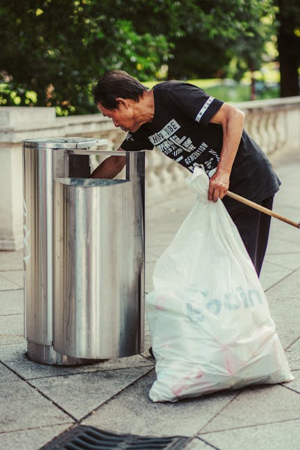 A middle-aged man with dark hair, wearing a black T-shirt with white text and dark trousers, is seen emptying a white plastic bag filled with rubbish into a public stainless steel waste bin. The bin has a shiny, reflective surface and is situated on a paved sidewalk, with the lid open as the man leans forward to deposit waste. In the background, there is a low stone wall and lush green trees, indicating an outdoor urban setting, possibly a park or a street with landscaped areas. The man appears focused on discarding waste properly, supporting independent rubbish disposal efforts. The scene is illuminated naturally, demonstrating a typical daytime environment suitable for outdoor waste collection and rubbish disposal services like those offered by houseclearancekingstonuponthames.co.uk, which often handle on-site clearance and private waste management tasks. The image emphasizes clean urban environments and individual responsibility in waste management, relevant for discussing alternative rubbish collection solutions outside regular municipal services.