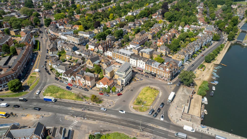 An aerial view of a town centre showing a mix of multi-storey and terraced buildings, with a busy intersection in the foreground and a river on the right side. The scene includes cars, vans, and trucks navigating the roads, with some parked along the streets and in designated parking areas. The buildings display a variety of brick and painted facades, with rooftops in dark and light colors, and some structures featuring flat roofs, pitched tiles, and decorative elements. Green trees and small patches of landscaped areas provide a contrast to the urban environment, while yachts and small boats are anchored along the riverbank. The overall environment appears lively and well-maintained, with clear weather and natural daylight illuminating the scene. This setting offers context for private waste management and alternative rubbish collection options in a suburban town, consistent with visual themes of urban rubbish removal and clearance services such as those provided by houseclearancekingstonuponthames.co.uk.