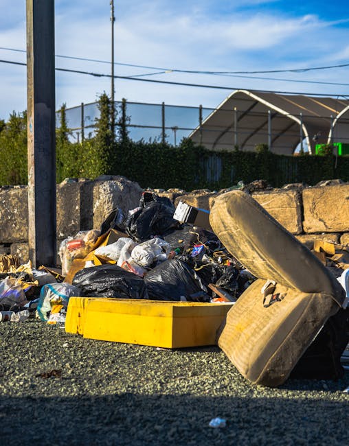 A pile of mixed waste materials is gathered on a paved surface outdoors, featuring various black and plastic garbage bags, some torn open revealing rubbish inside. There are several larger items, including a worn, beige car tire leaning against the pile and a yellow plastic container or bin partially visible among the refuse. In the background, a low stone wall made of irregularly shaped, rough-hewn stones separates the waste area from a green hedge and a large, metallic-framed canopy or industrial structure with a curved roof, set against a blue sky with scattered clouds. Overhead power lines run across the scene, suggesting an urban or semi-industrial environment. The lighting indicates broad daylight, casting sharp shadows and highlighting the textures and colors of the waste and surroundings, illustrating a typical scene requiring rubbish removal services that handle alternative waste disposal at or near private property or commercial sites.