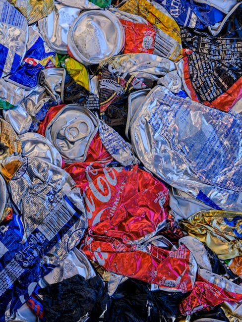 A close-up view of a pile of crushed and flattened aluminium beverage cans, primarily made of silver-colored aluminum with some featuring blue, red, yellow, and black printed labels and logos. The cans vary in size and design, with some showing recognizable branding such as Coca-Cola. They are crumpled and stacked together, occupying the entire frame, with visible textures of metallic surface and printed paper labels. The background is not visible, indicating a dense accumulation of waste material. This scene is representative of recyclable aluminium waste, often collected for recycling and disposal through private waste management services such as houseclearancekingstonuponthames.co.uk, which handle non-standard rubbish removal at a household or commercial level. The lighting highlights the reflective metallic surfaces and the vibrant colors of the logos and labels on the cans, emphasizing their material composition and the need for proper disposal or recycling as part of alternative waste handling methods.
