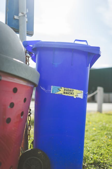 A pile of mixed waste materials is gathered on a paved surface outdoors, featuring various black and plastic garbage bags, some torn open revealing rubbish inside. There are several larger items, including a worn, beige car tire leaning against the pile and a yellow plastic container or bin partially visible among the refuse. In the background, a low stone wall made of irregularly shaped, rough-hewn stones separates the waste area from a green hedge and a large, metallic-framed canopy or industrial structure with a curved roof, set against a blue sky with scattered clouds. Overhead power lines run across the scene, suggesting an urban or semi-industrial environment. The lighting indicates broad daylight, casting sharp shadows and highlighting the textures and colors of the waste and surroundings, illustrating a typical scene requiring rubbish removal services that handle alternative waste disposal at or near private property or commercial sites.