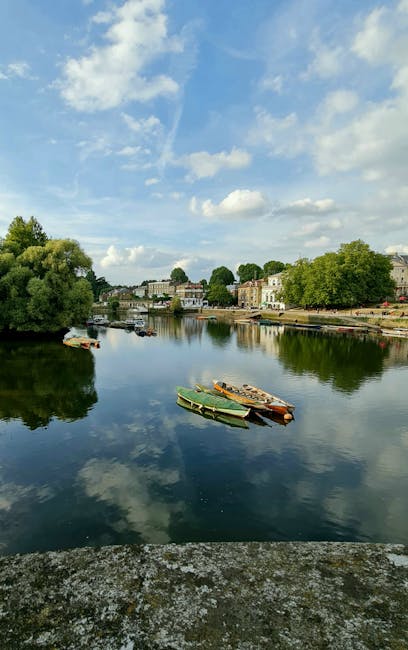 A calm river scene with a reflective water surface showing partly cloudy blue sky and scattered white clouds overhead. In the foreground, a stone or concrete edge lines the riverbank. Several small boats, including a green rowboat and a couple of orange and brown boats, are moored together near the shore on the water's surface. Across the river, on the far bank, there are residential and commercial buildings with light-colored facades and tiled roofs, surrounded by lush green trees and foliage. The overall scene suggests a peaceful, semi-urban setting, possibly in a historic or conservation area, with the river serving as a central feature. This quiet environment aligns with the context of private waste management or alternative rubbish removal services, such as on-site clearance or independent collection options often used in protected or regulated areas.