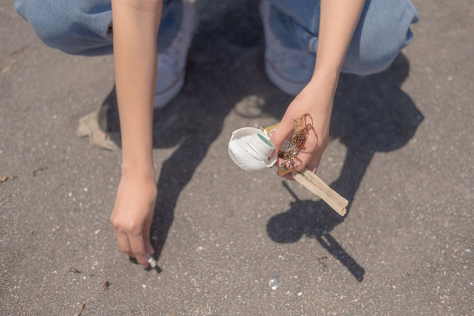 A person wearing blue jeans and white sneakers is crouching on an asphalt surface, holding a disposable white foam cup with a green stripe and a piece of wooden stick, with remnants of food or other debris attached to it. The person's arm and hand are visible, positioned centrally in the image, with a shadow cast on the ground. The background shows a ground surface with small stones and dirt. This scene highlights the importance of proper waste disposal and can be associated with rubbish removal services such as those offered by houseclearancekingstonuponthames.co.uk for managing litter and waste in outdoor urban environments.