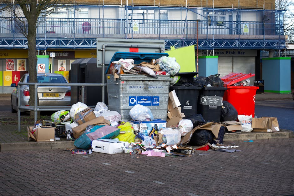 An outdoor scene in a commercial or residential area shows a large pile of mixed waste and rubbish overflowing from several open-top bins and disposal containers. The collection includes various packaging materials, cardboard boxes, plastic bags, paper, and loose debris. The waste is visibly spilling onto the surrounding paved ground, with some items partially collapsed or crumpled. Behind the waste, there are parked cars, and in the background, a building under scaffolding with blue protective netting indicating ongoing construction or renovation work. The area is illuminated by natural daylight, casting slight shadows around the rubbish and vehicles. The scene captures a typical instance of potentially unmanaged rubbish accumulation, reflecting scenarios where private waste handling or alternative collection services like those provided by houseclearancekingstonuponthames.co.uk could be relevant for proper waste removal and disposal.