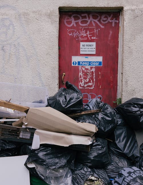 A pile of black rubbish bags and discarded packaging materials, including a cardboard box and a piece of foam, are stacked against a wall near a red metal door covered with graffiti. The door has a small white sign that states 'No Money' and a blue and white sticker reading 'Keep Clear.' The setting appears to be an outdoor urban area, with the rubbish lying directly on a concrete surface beside a textured, off-white wall. The scene suggests an instance of unmanaged waste awaiting collection or disposal, consistent with services offered by companies like houseclearancekingstonuponthames.co.uk for private rubbish removal or on-site clearance, in contrast to municipal waste collection. The lighting is natural, likely daylight, illuminating the area evenly and emphasizing the textures of the bags, the rough wall surface, and the graffiti-covered door. Overall, the image captures a typical example of unmanaged street-level waste accumulation in an urban environment that requires professional rubbish removal services for effective clearance.