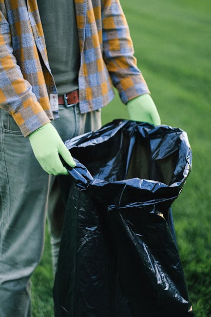 An outdoor scene in a commercial or residential area shows a large pile of mixed waste and rubbish overflowing from several open-top bins and disposal containers. The collection includes various packaging materials, cardboard boxes, plastic bags, paper, and loose debris. The waste is visibly spilling onto the surrounding paved ground, with some items partially collapsed or crumpled. Behind the waste, there are parked cars, and in the background, a building under scaffolding with blue protective netting indicating ongoing construction or renovation work. The area is illuminated by natural daylight, casting slight shadows around the rubbish and vehicles. The scene captures a typical instance of potentially unmanaged rubbish accumulation, reflecting scenarios where private waste handling or alternative collection services like those provided by houseclearancekingstonuponthames.co.uk could be relevant for proper waste removal and disposal.