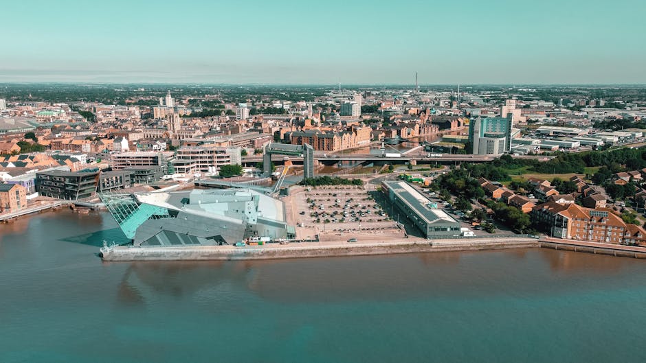 An aerial view of a cityscape featuring a modern building with angular, metallic, and glass facades situated along a riverbank, with a parking lot adjacent to it. Behind the building, there are extended areas of open land, further urban development, and a densely built environment comprising various low-rise and mid-rise structures. The river in the foreground reflects the structures and sky, while the background reveals an expansive skyline with numerous commercial and residential buildings under a pale, slightly overcast sky. This scene exemplifies urban infrastructure, where private waste handling or on-site clearance operations could be visually associated with the surrounding environment, fitting into a context of alternative rubbish removal services that focus on independent collection and management of waste.