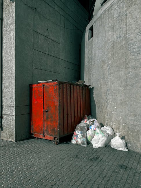A large, rusty red metal skip with a corrugated surface, situated against a dark gray wall and a light stone wall in an outdoor urban setting. The skip has visible signs of wear, including chipped paint and surface rust, indicating frequent use for waste collection. Several white and transparent plastic rubbish bags filled with various waste materials are placed on the ground beside the skip, some leaning against it. The ground features dark gray paving stones arranged in a grid pattern, with the overall environment suggesting a designated waste disposal area for private or commercial rubbish removal. The scene is lit by natural daylight, casting soft shadows and emphasizing the textures of the metal, plastic, and surrounding walls, aligning with practices of independent collection or on-site clearance managed by waste removal services like houseclearancekingstonuponthames.co.uk.